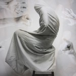 A digital image of a figure wrapped in a white fabric with hand pointing at it in the background