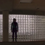 Still image of sihouletted woman standing in front of a glass block wall.